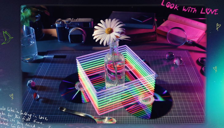 A daisy in a bottle surrounded by colorful light filters, CDs, a camera, magnifying glass, and books. Text reads "Look with Love" in the upper corner.