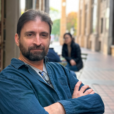 A man with a beard and crossed arms stands on a street with blurred people and buildings in the background.