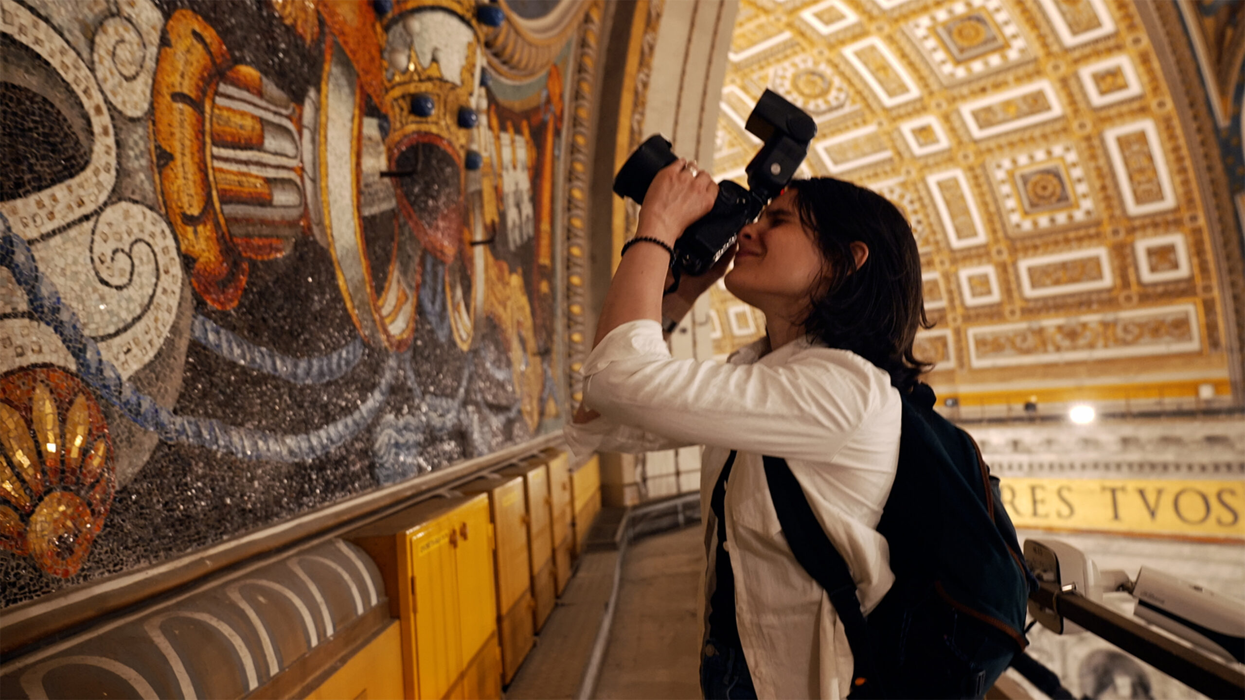 Person photographing a detailed mosaic on a cathedral wall with an ornate ceiling overhead.