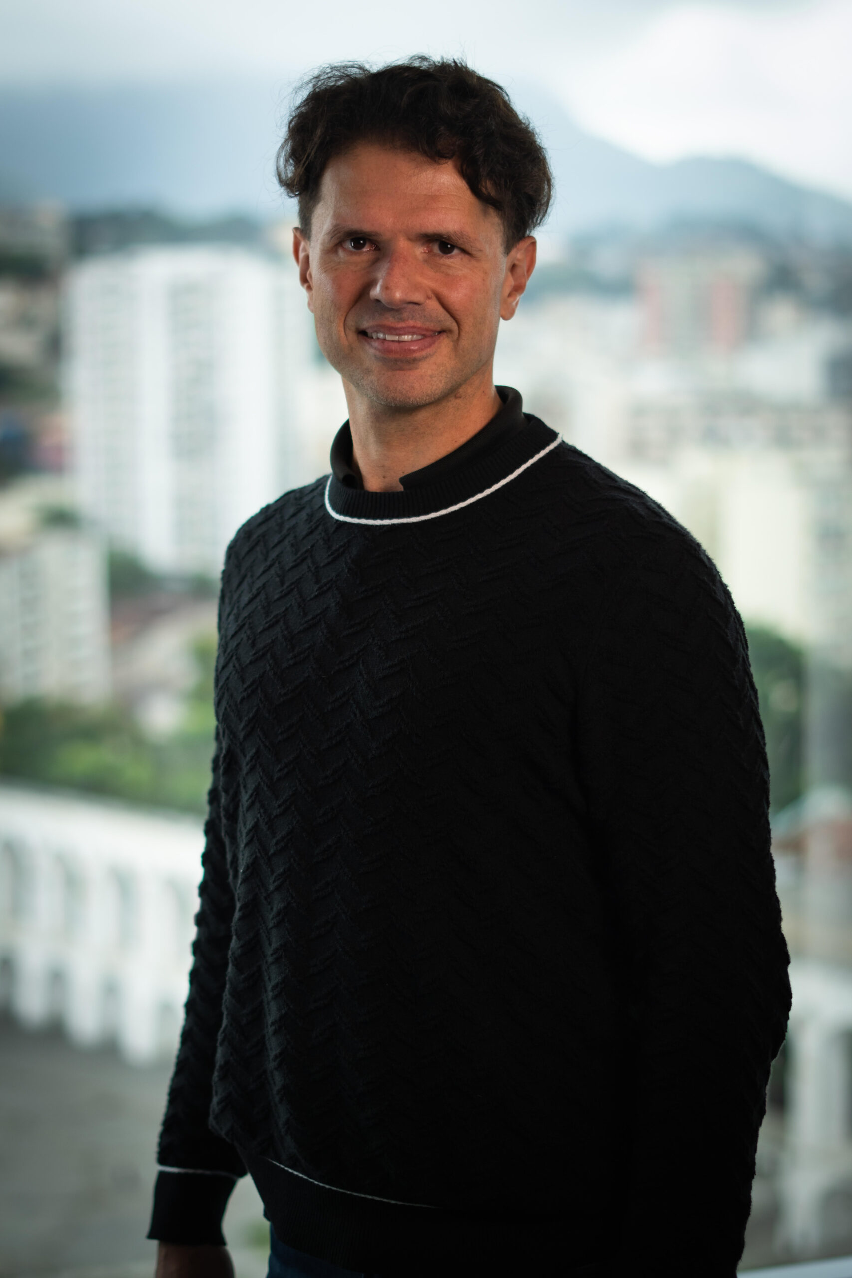 A person with short, dark hair wearing a black sweater stands indoors with a cityscape visible through the large window behind them.