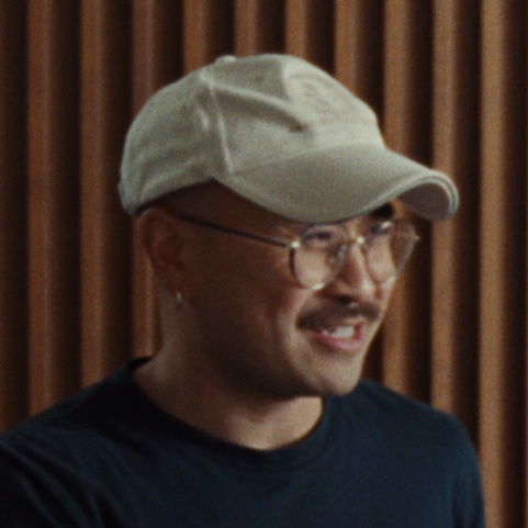 Person wearing a white cap and glasses, smiling, against a backdrop of vertical wooden panels.