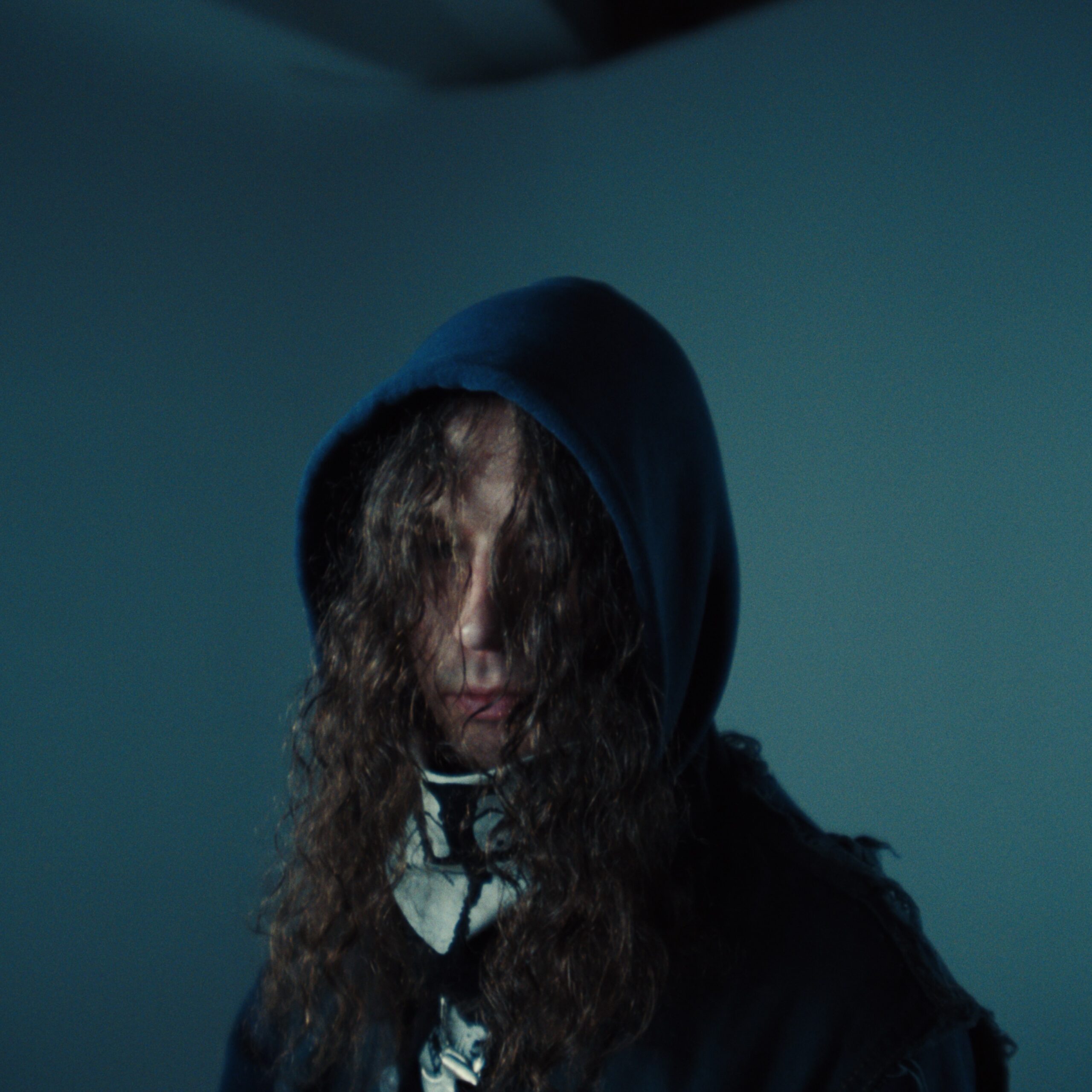 Person wearing a dark hooded garment and scarf, with long curly hair partially obscuring their face, standing against a plain wall.