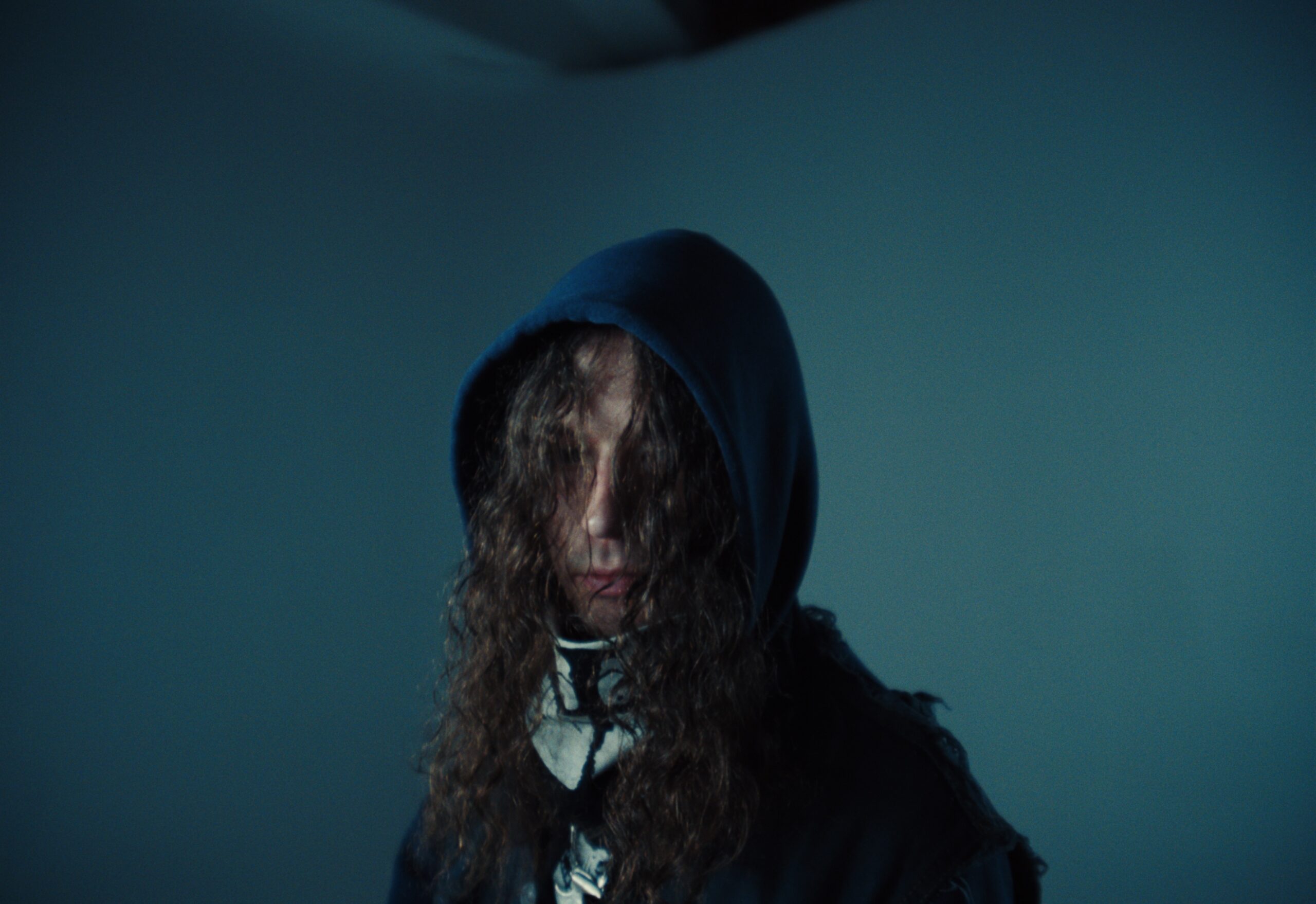 Person with long curly hair wearing a dark hooded garment, standing against a plain background.