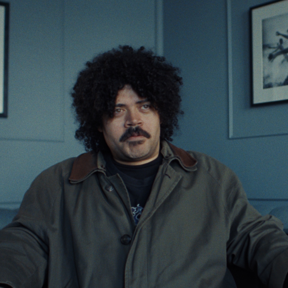 Man with curly hair and a mustache sits in a room with blue walls and framed pictures.