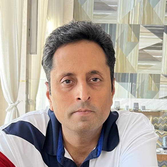 A man with short dark hair and a calm expression is wearing a white shirt with blue and black accents. He is indoors, with patterned walls and white curtains in the background.