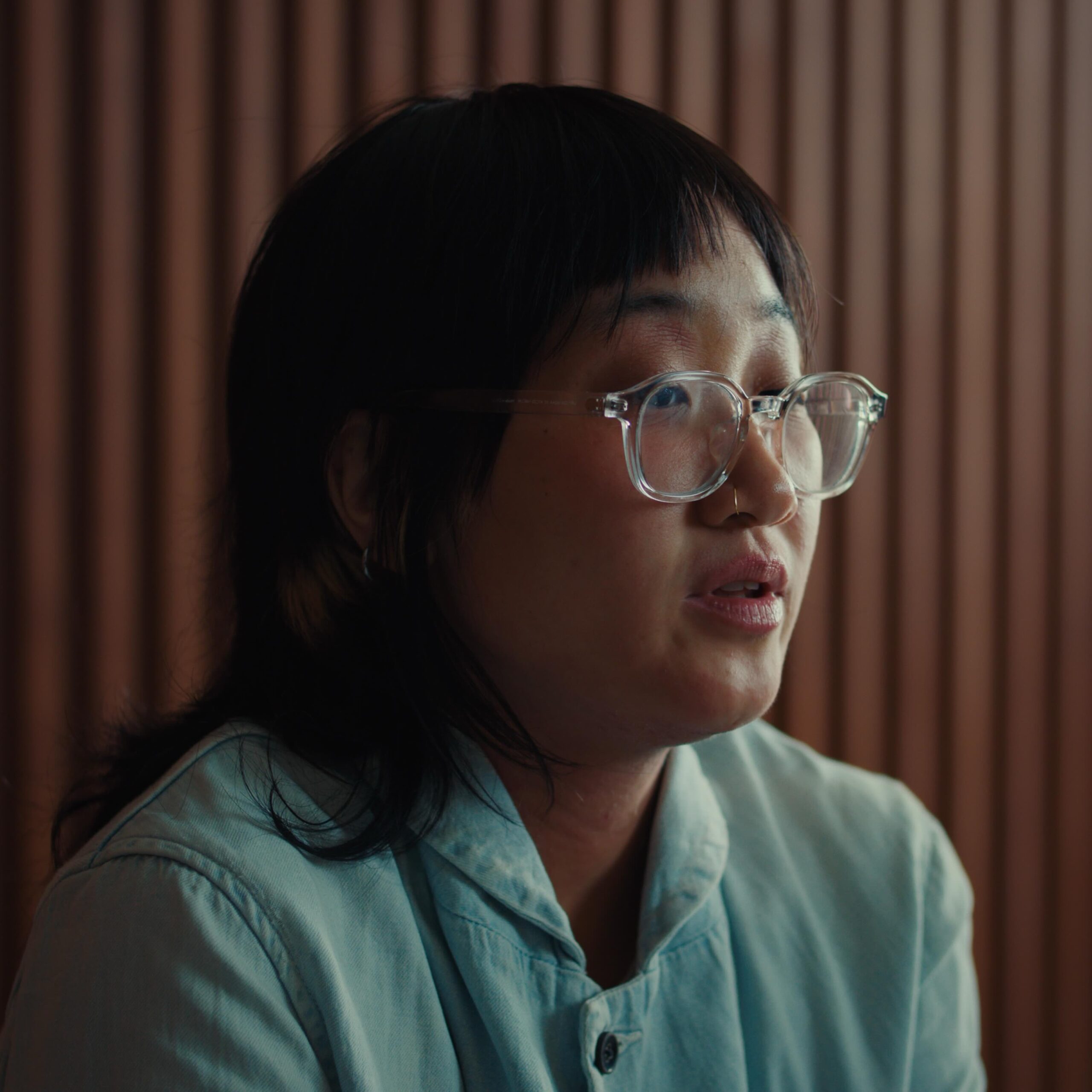 A person with glasses and long black hair wearing a light blue shirt sits in front of a wooden, vertical panel backdrop.