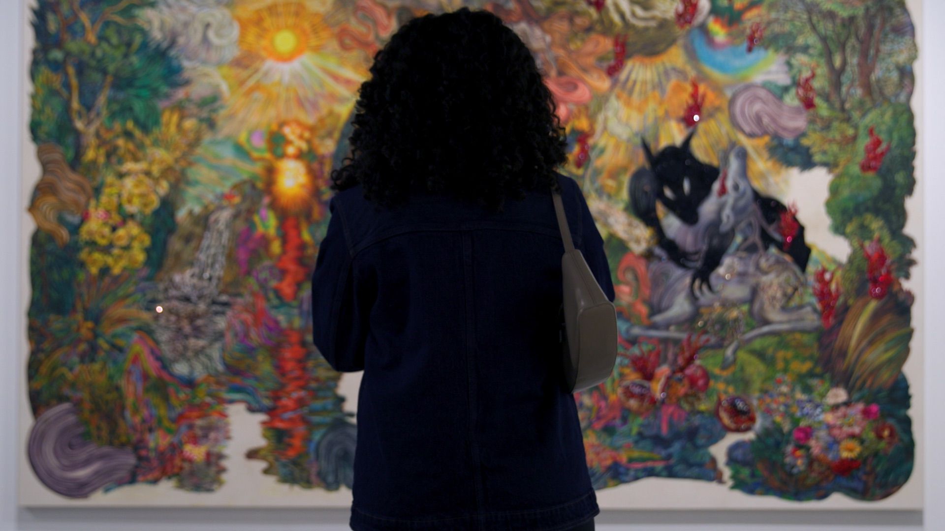 Person with curly hair observes a colorful, abstract painting in a museum or gallery setting.