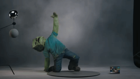 A person in a green costume poses dramatically on one knee in a studio. There is a lighting setup and a color chart nearby.
