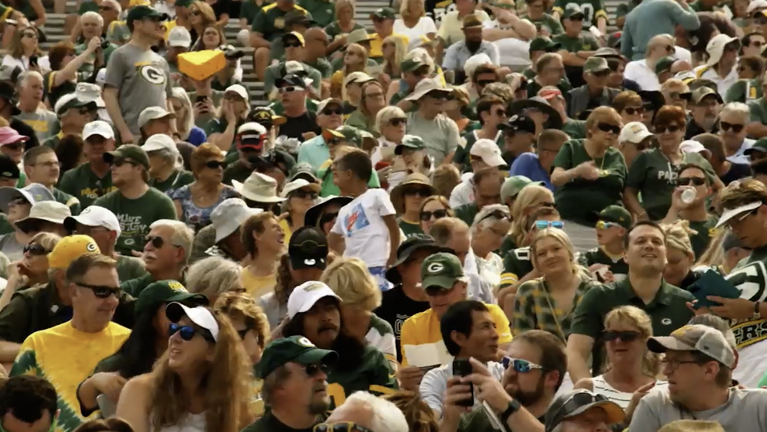 A large crowd of people, many wearing green and yellow sports apparel and hats, sit closely together in stadium seating, watching an event.