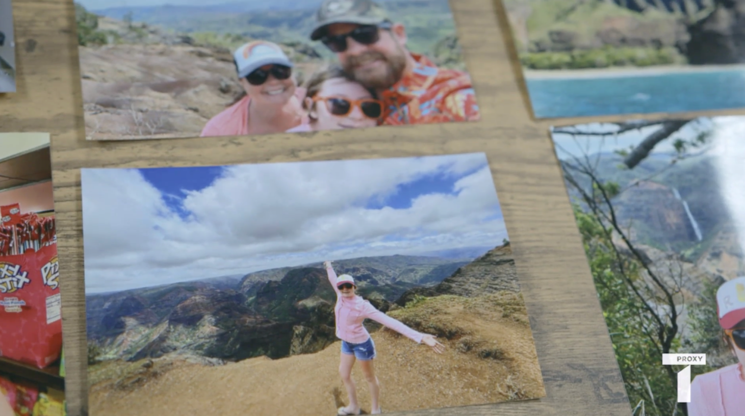 A photograph on a wooden surface shows a woman in a pink shirt and hat posing with arms outstretched at a scenic mountain overlook. Other photos of outdoor scenes surround it.