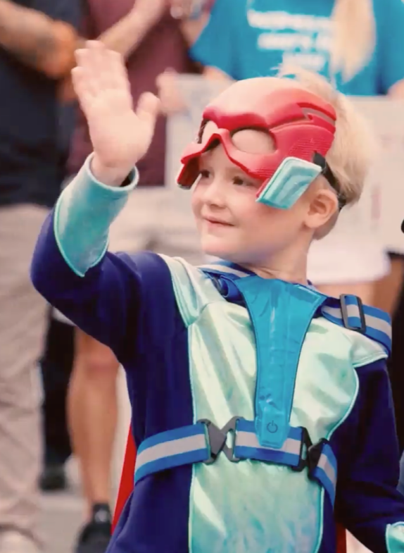 A young child in a colorful superhero costume and red mask waves while standing outdoors among a crowd.