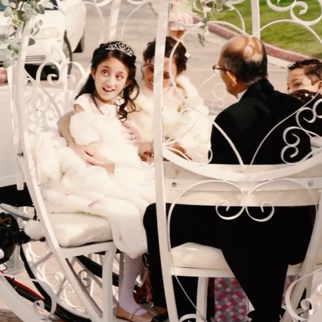 A girl in a white dress and tiara sits in a decorative, flower-adorned carriage with three other people on a street.