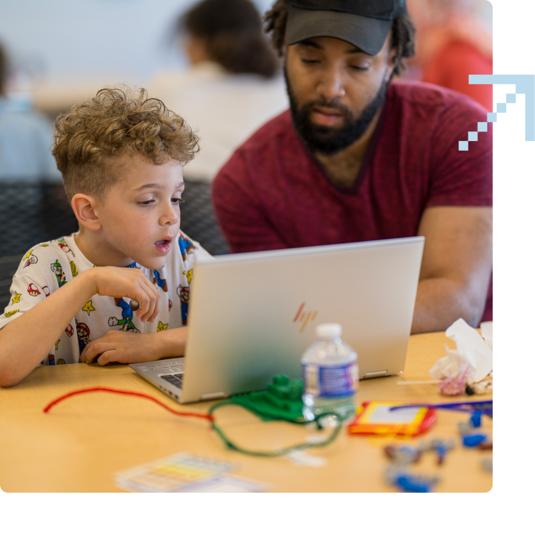 A man and a boy sit at a table looking at a laptop screen together. Various objects and a water bottle are on the table in front of them.