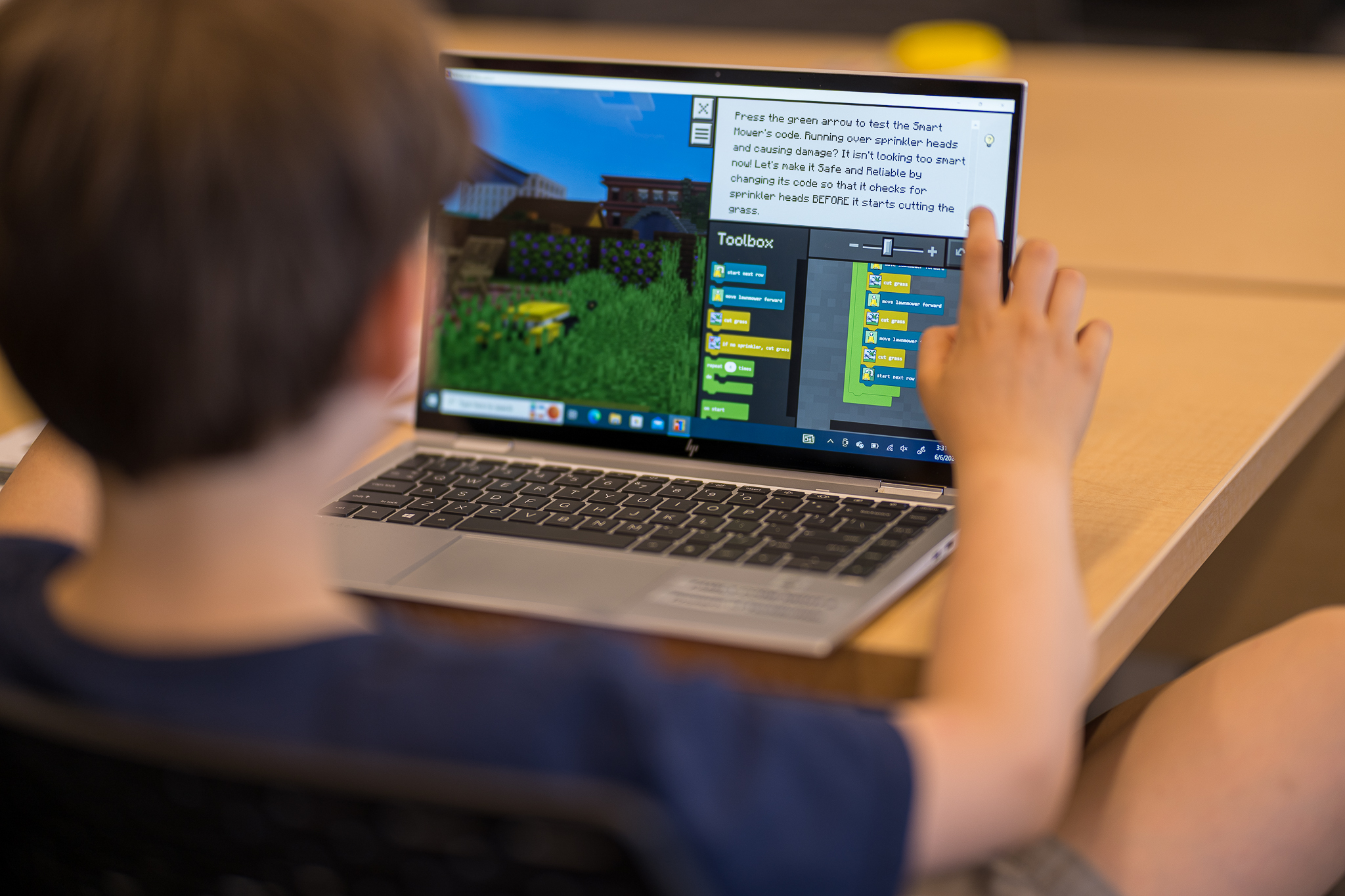 A child uses a laptop to work on a coding project in Minecraft, with visual programming blocks displayed on the screen.