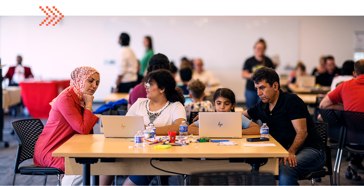 Four people sit at a table using laptops and talking, with other groups working in the background in a brightly lit room.