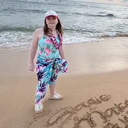 A person in a floral outfit stands on a sandy beach near ocean waves, with writing in the sand at their feet.
