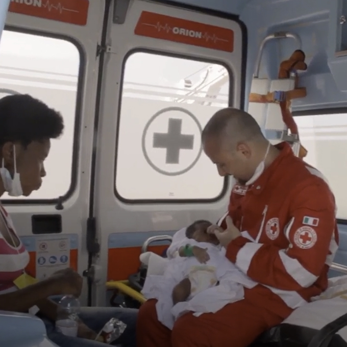 A Red Cross worker cares for an infant inside an ambulance while a woman sits nearby wearing a mask, with medical supplies visible in the background.