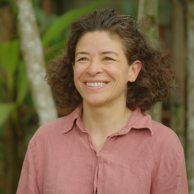 A woman with curly brown hair and a nose ring, wearing a pink button-up shirt, smiles while standing outdoors with trees and greenery in the background.