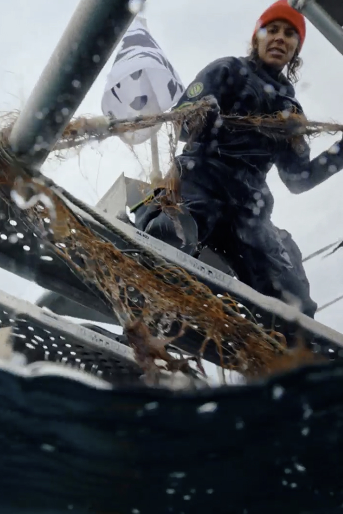 A person in a wetsuit and orange beanie stands on a metal structure above water, removing tangled fishing net debris; a flag with a panda logo is visible.