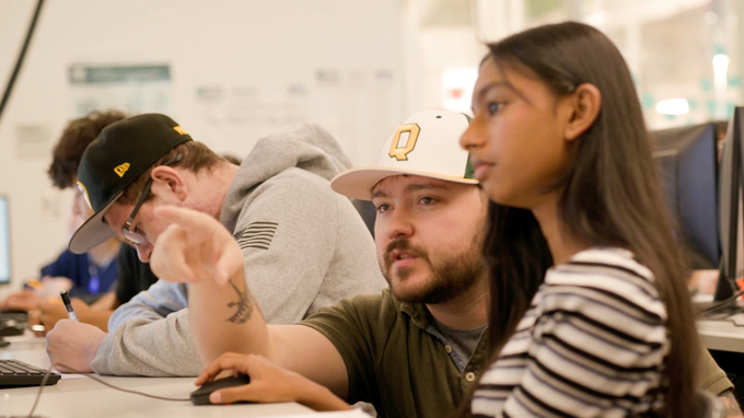 Two people sit at a desk with computers; one points at the screen while the other looks on attentively. Others work in the background.