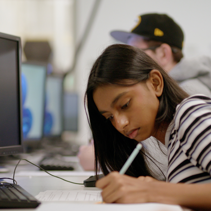 A student with long dark hair writes on paper at a desk with computers, while another person sits in the background.