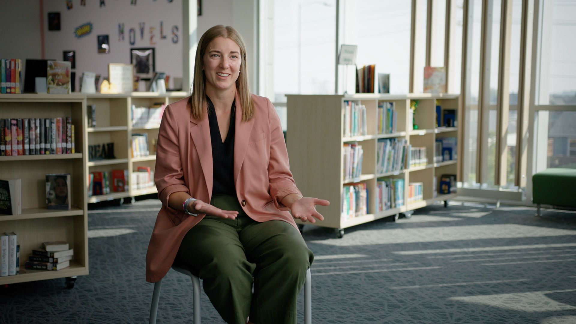 A woman in a pink blazer and green pants sits on a chair in a library, gesturing with her hands and smiling. Bookshelves and large windows are visible in the background.