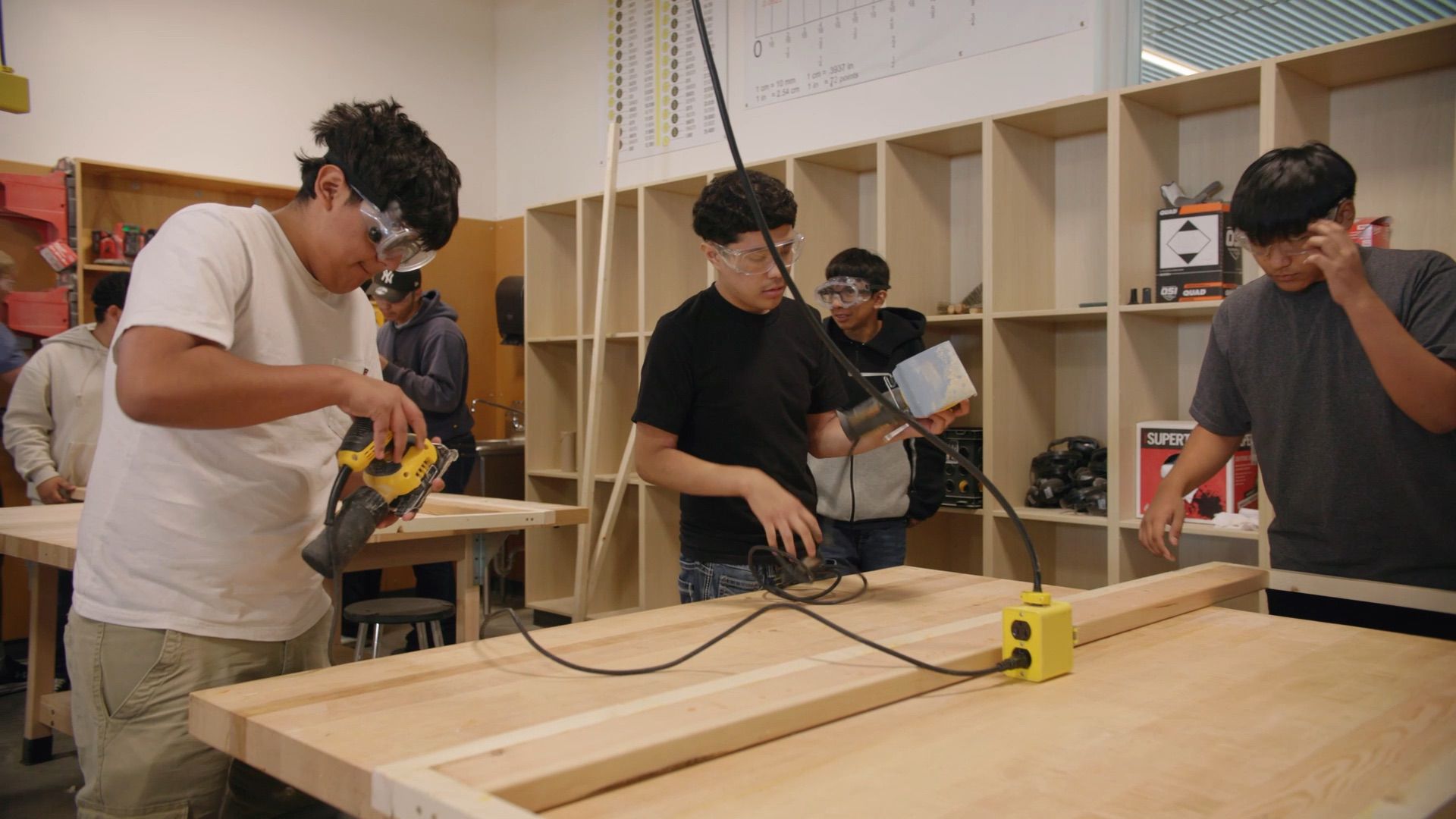 Four students wearing safety goggles work with tools and wood at a workshop table in a classroom setting.