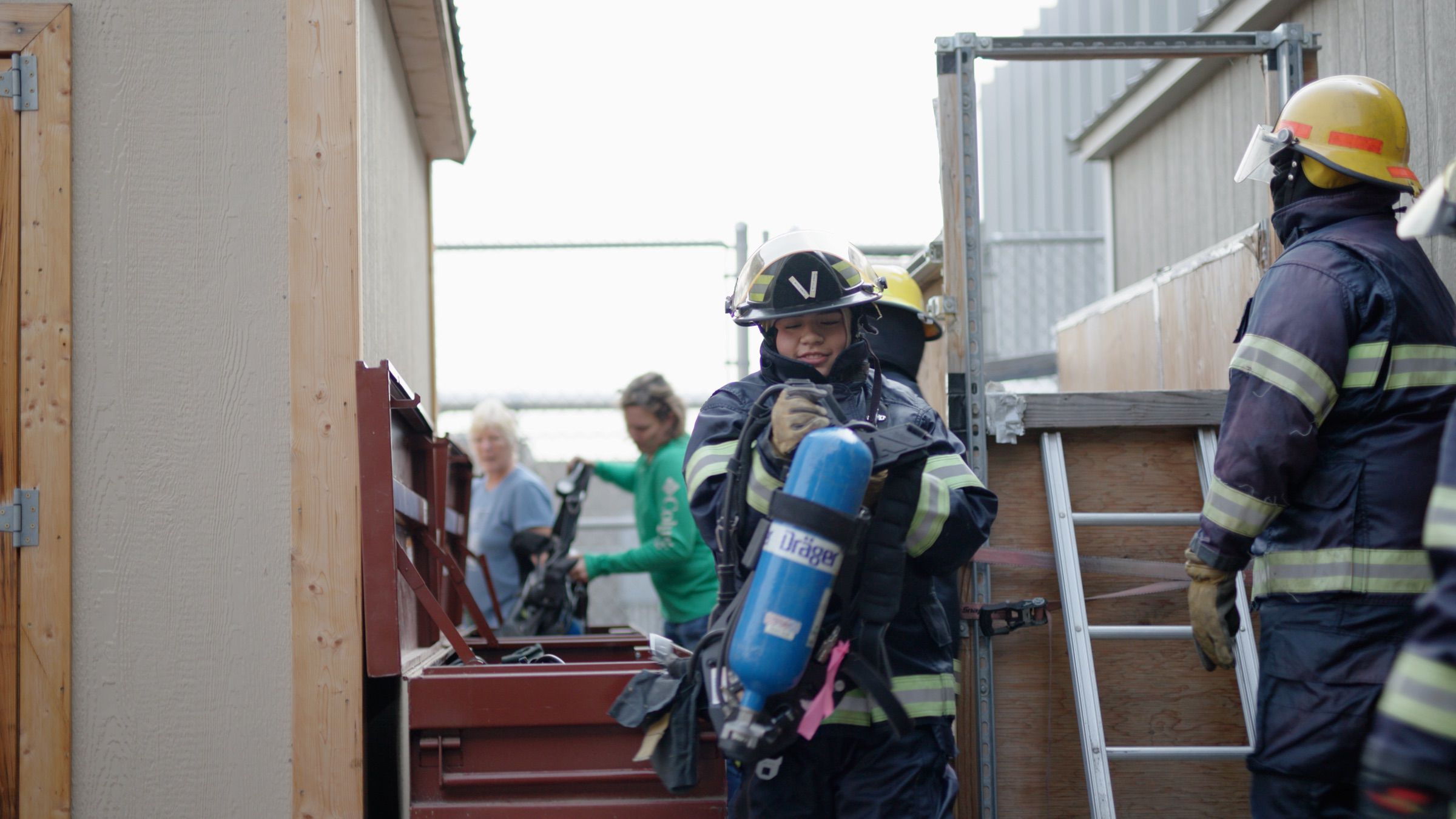 Firefighters in full gear walk between buildings, with one carrying a breathing apparatus. Several people are in the background near equipment and a ladder.