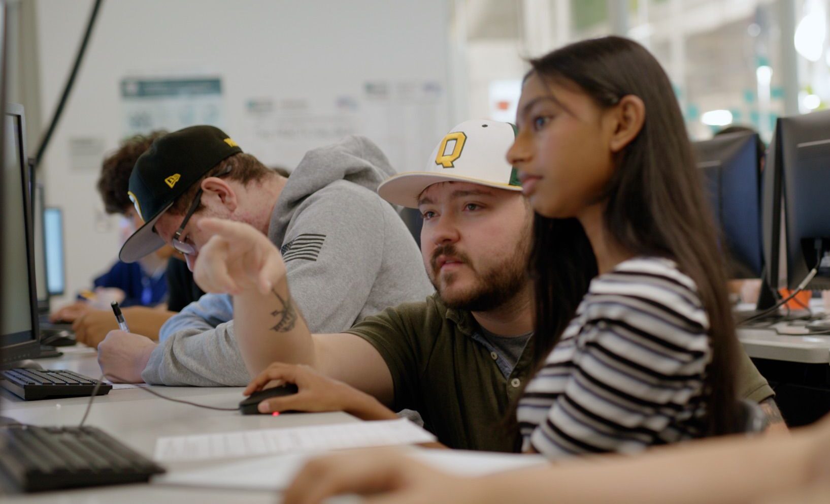 Two students sit at a computer in a classroom; one points at the screen while the other follows, with other students working in the background.
