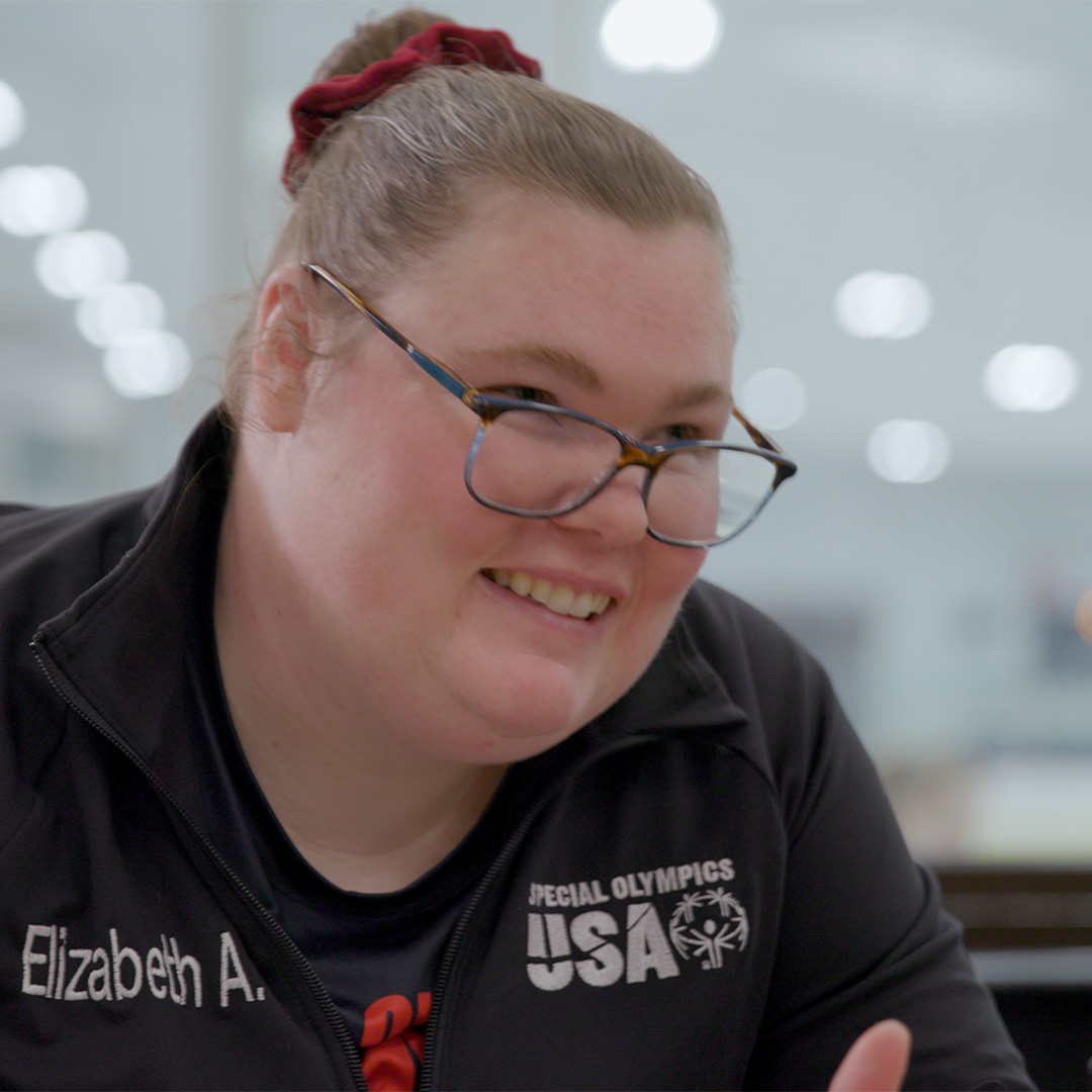Una mujer con gafas y una dona roja en el cabello sonríe mientras usa una chaqueta negra de patinaje de Special Olympics USA. Su nombre, “Elizabeth A.”, es visible en la chaqueta. El fondo está suavemente desenfocado.  