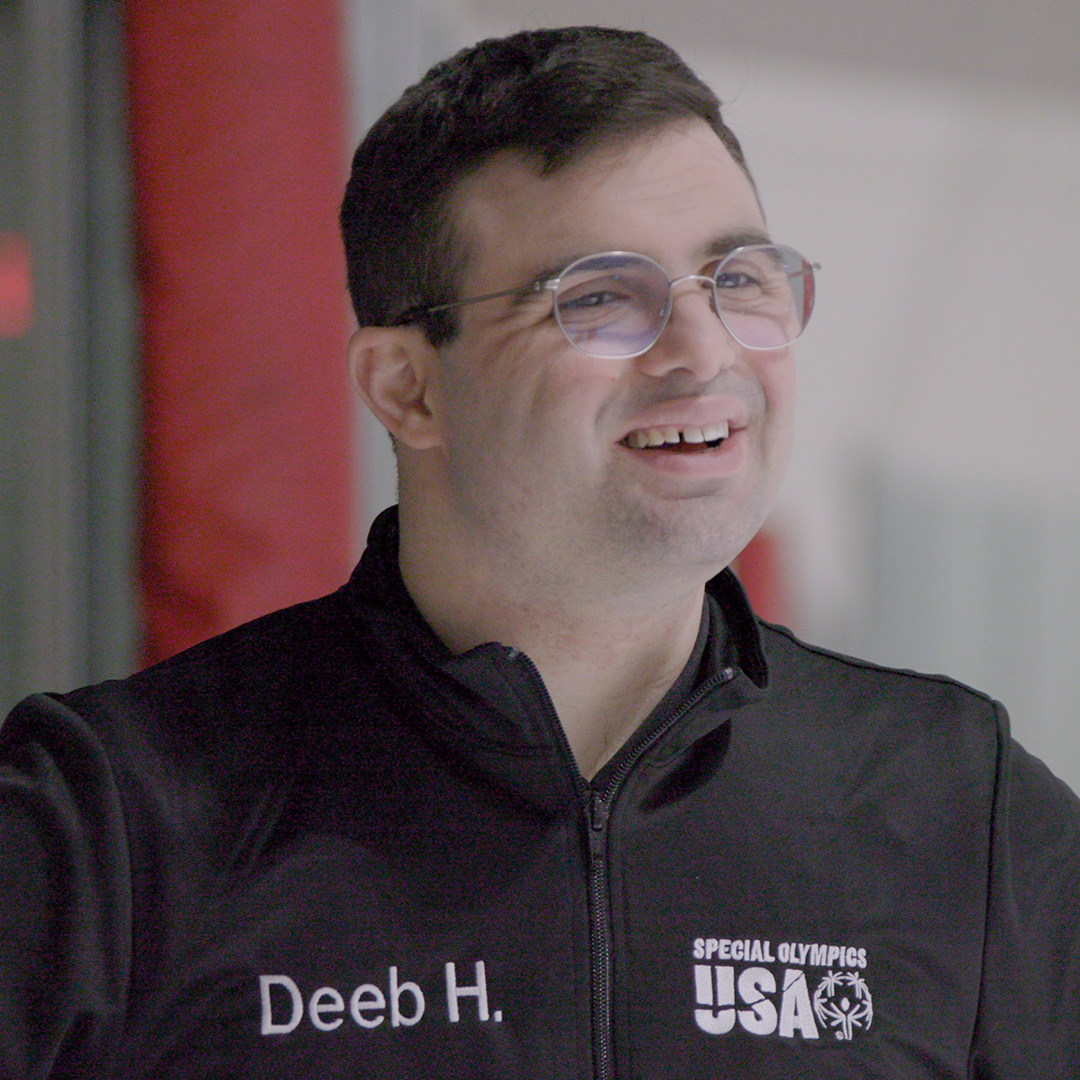 Un hombre de cabello oscuro corto y gafas sonríe mientras usa una chaqueta negra con los logotipos de “Deeb H.” y de Special Olympics USA bordados, celebrando su amor por el patinaje.