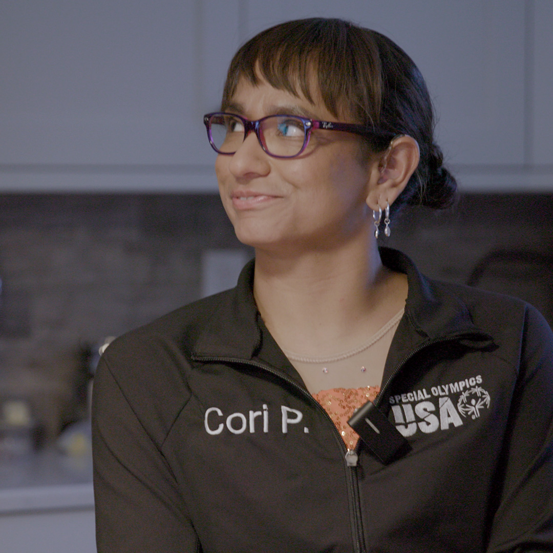 Una mujer de cabello oscuro recogido en un chongo, que lleva gafas, aretes y una chaqueta negra de patinaje de Special Olympics USA con el nombre “Cori P.” bordado, está sentada en interiores y sonríe levemente mientras mira hacia su izquierda.