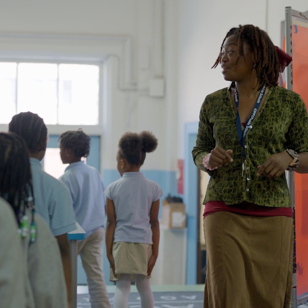 A teacher stands and smiles while watching young students in school uniforms walking away down a hallway in a brightly lit school building.