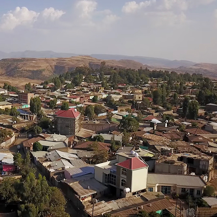 Aerial view of a town with clusters of houses, some with red roofs, surrounded by trees and hills under a partly cloudy sky.