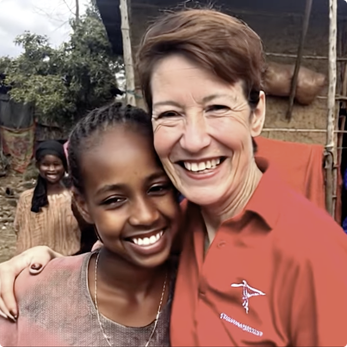 A smiling woman in a red shirt hugs a smiling young girl outdoors, with another girl standing in the background near a rustic building.