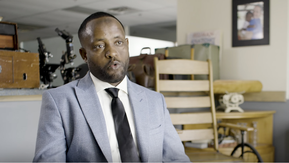 A man in a light blue suit and black tie sits indoors, speaking. Behind him are a wooden chair, vintage items, and framed photos on the wall. The setting appears to be an office or interview space.