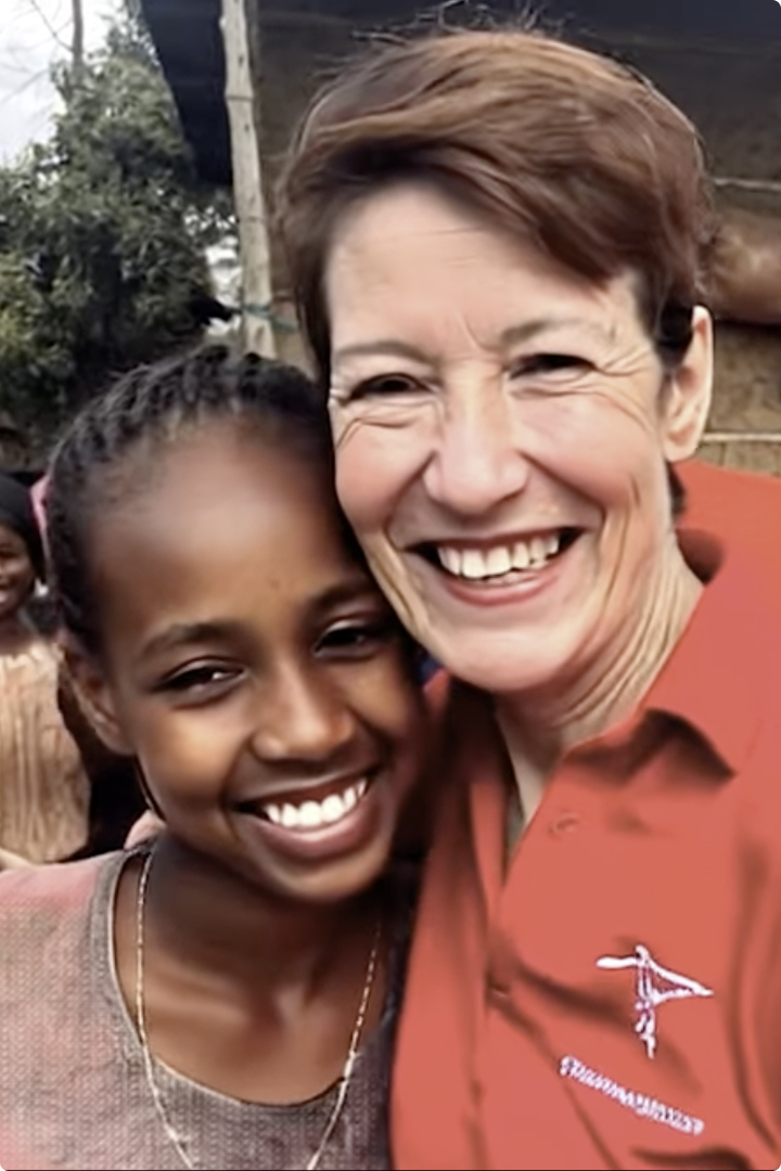 A smiling woman in a red polo shirt hugs a smiling young girl outdoors. Several people and a building appear blurred in the background.