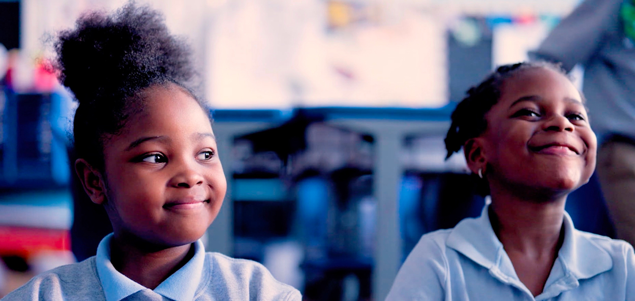 Two young girls in school uniforms sit side by side, smiling and looking in different directions, with a classroom background softly out of focus.