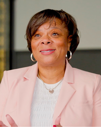 A woman wearing a light pink blazer and white blouse smiles, gesturing with her hands. She has short brown hair, hoop earrings, and a heart-shaped necklace, standing against a blurred, neutral background.