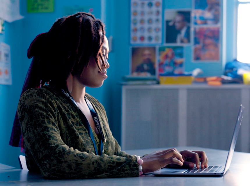 A person with long, dark hair and a patterned green top types on a laptop at a desk in a brightly lit room decorated with posters and blue walls.