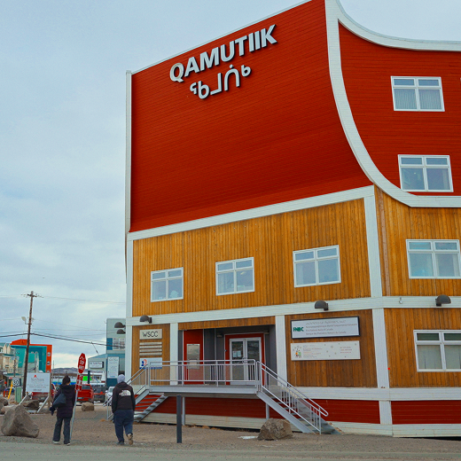 A red and yellow building labeled QAMUTIK with Inuktitut script beneath the sign; two people stand outside on a gravel lot under a cloudy sky.