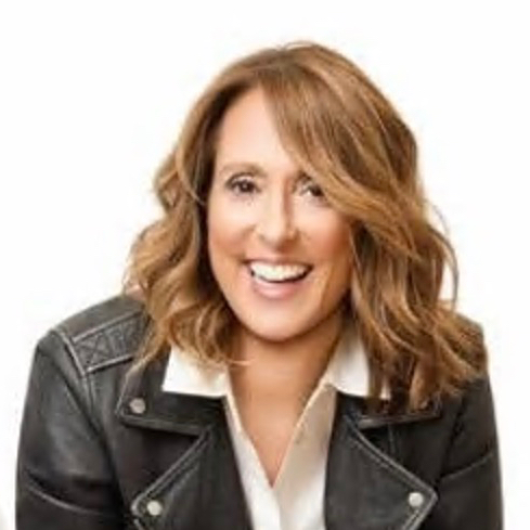 A woman with wavy, shoulder-length light brown hair smiles at the camera. She is wearing a black leather jacket over a white collared shirt against a plain white background.