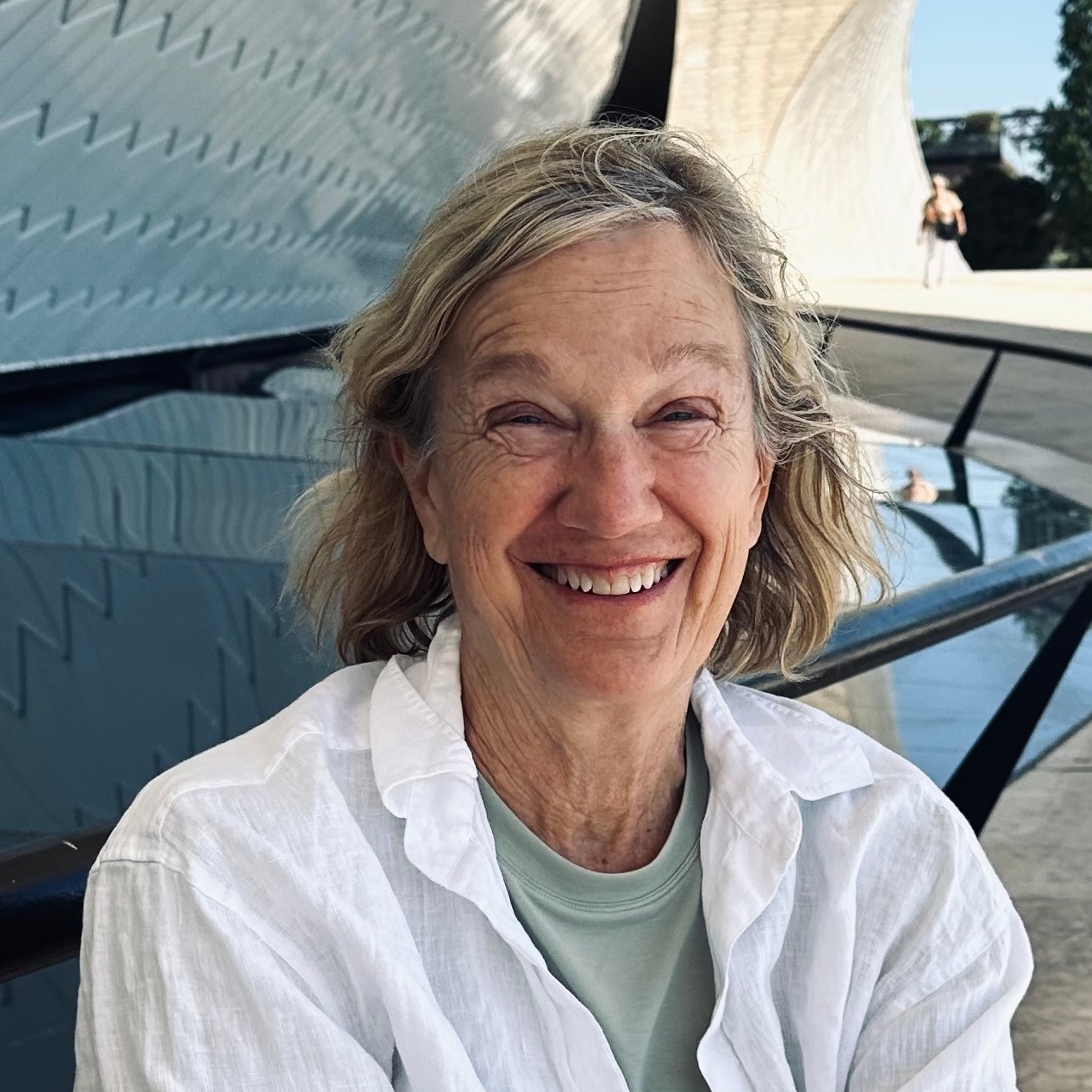 An older woman with shoulder-length gray hair smiles warmly, wearing a white shirt over a light green top. She is outdoors near a modern, curved architectural structure with sunlight and a person in the background.