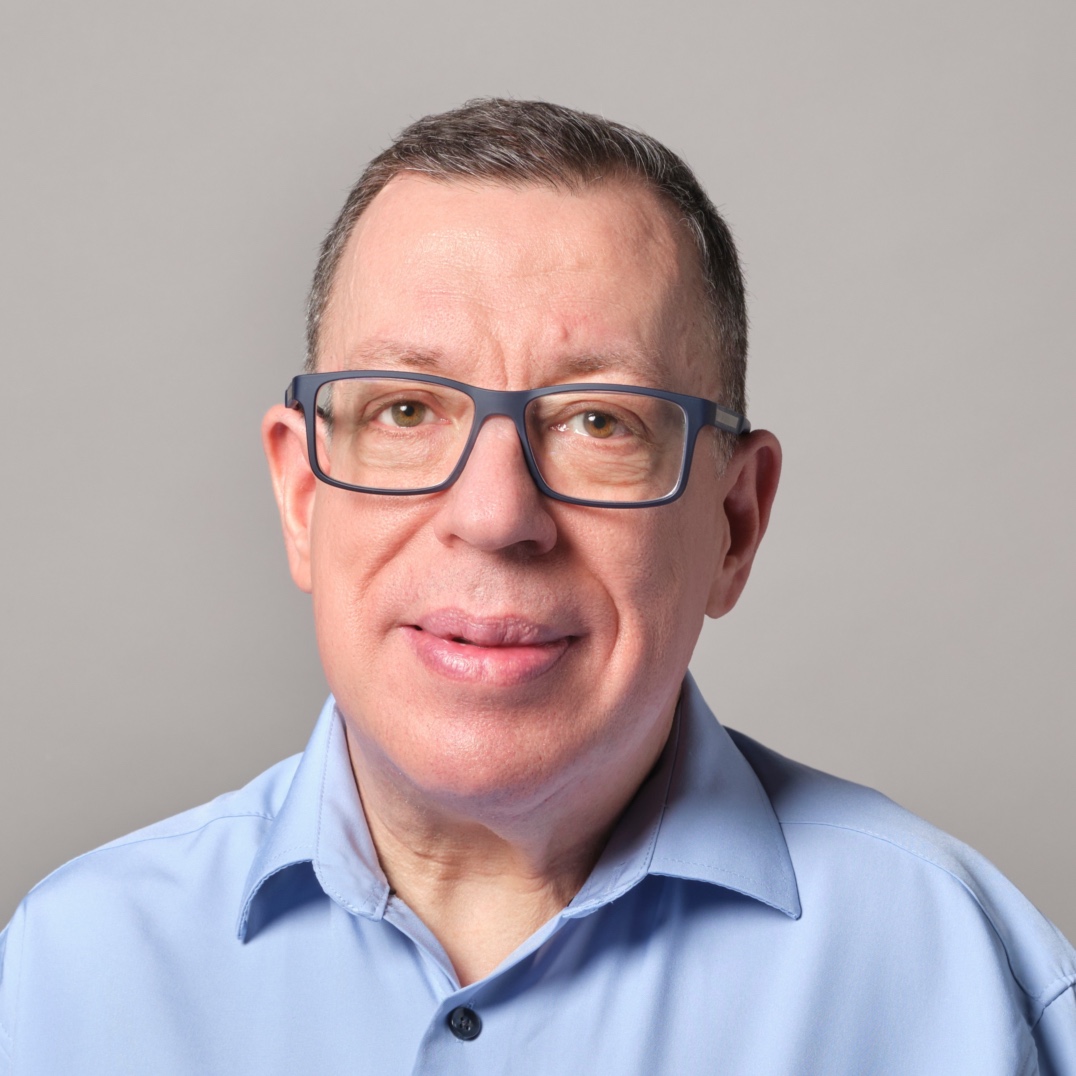 A middle-aged man with short brown hair, glasses, and a light blue collared shirt is smiling gently at the camera against a plain gray background.