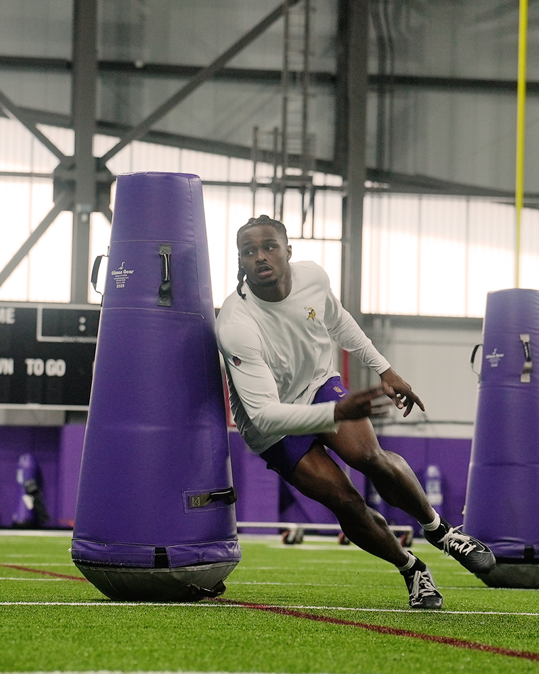 A football player in a white shirt and purple shorts runs around a tall purple tackling dummy during an indoor practice on artificial turf.