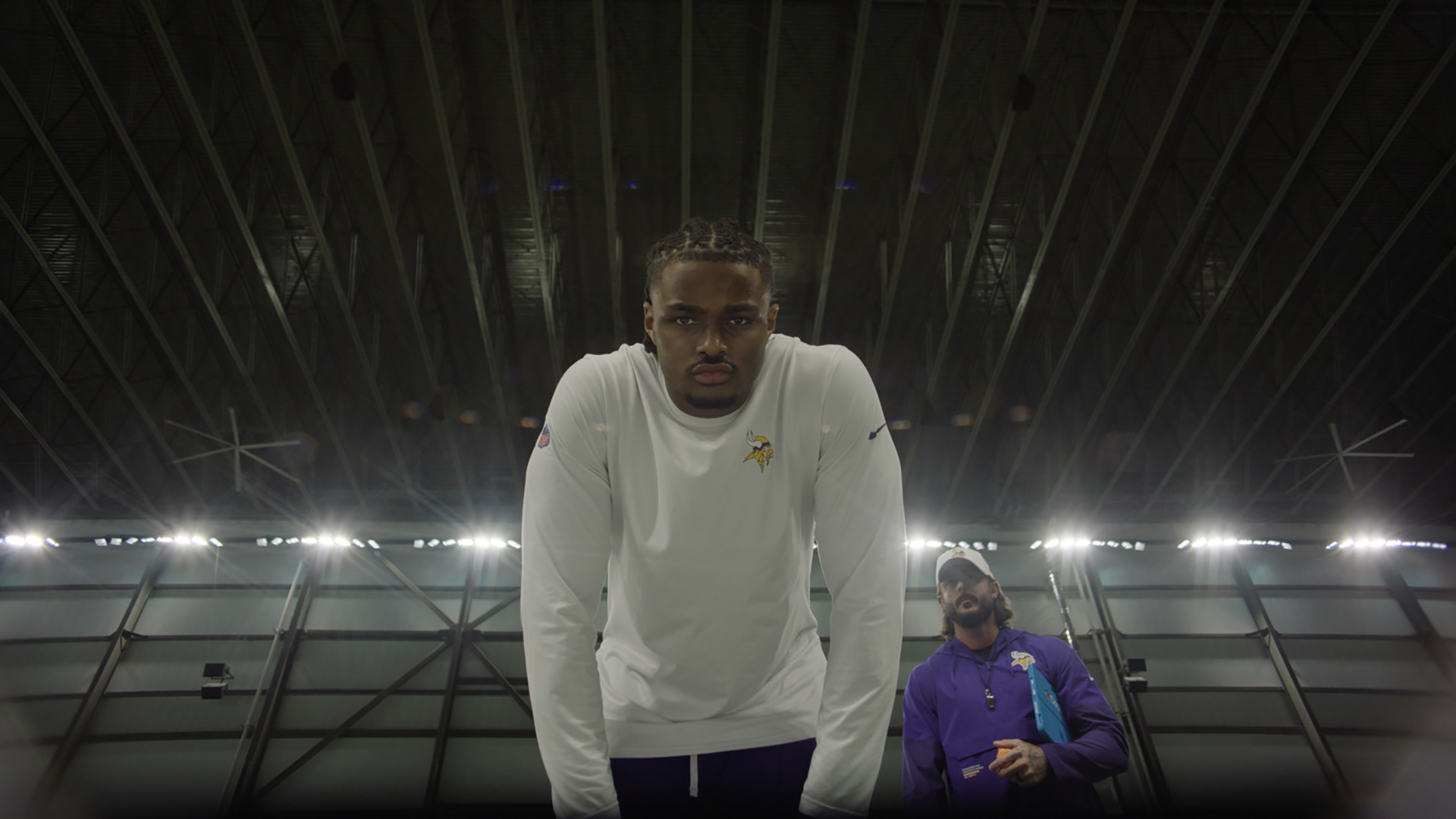 A football player in a white Minnesota Vikings shirt leans forward, looking intensely ahead, with another person in a purple jacket standing behind him inside a brightly lit indoor stadium.