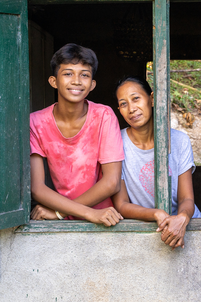Two smiling people, a teen boy and woman, lean out of a rustic window with weathered green wooden shutters on a concrete wall.