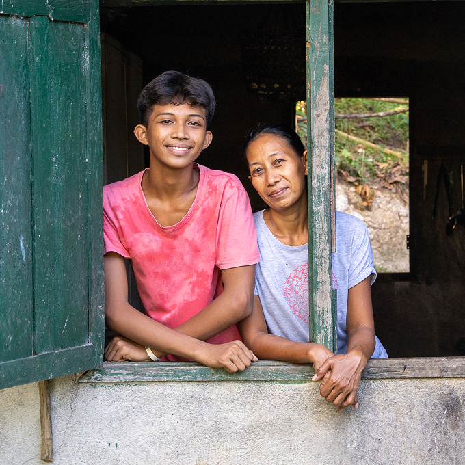 Two smiling people, a teen boy and woman, lean out of a rustic window with weathered green wooden shutters on a concrete wall.