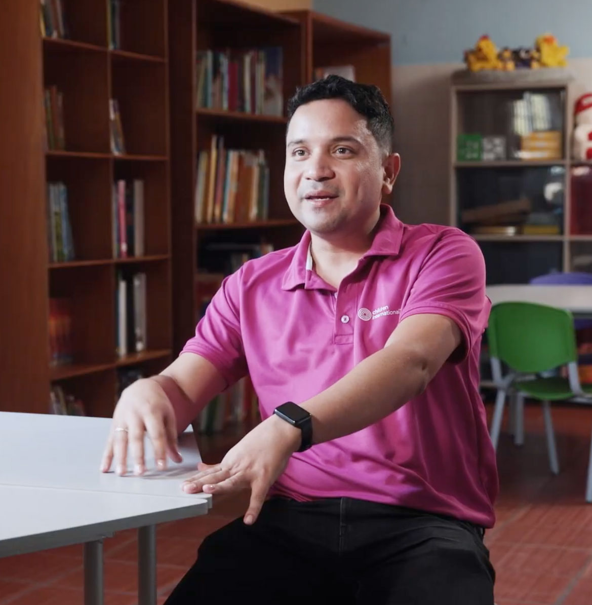 Man in pink polo shirt sitting at white desk in office with bookshelves in background