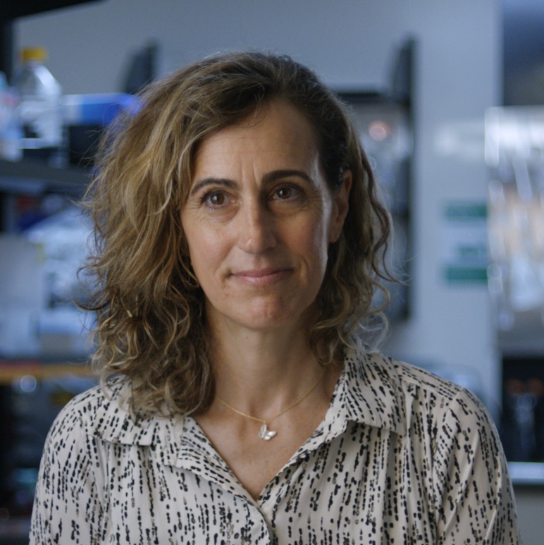 Woman with wavy brown hair wearing a white patterned blouse standing in a scientific laboratory with equipment on shelves behind her.